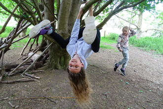 Child smiling to camera, hanging upside down from tree branch with long hair dangling. Another child sits on a rope swing looking on.  