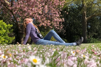 Woman sitting on the grass in a park, on a sunny day, surrounded by nature including a tree in blossom and daisies on the ground