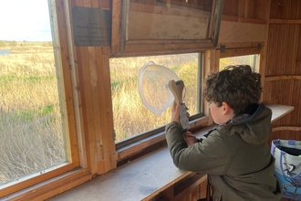 A boy sits on a bench looking out of the window of a bird hide over reedbeds. He holds a smart phone attached to a bowl to amplify the sounds