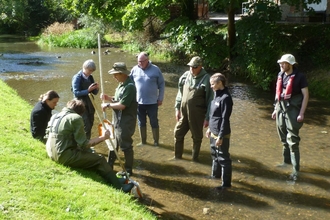 A group of 8 people in waders and wellington boots standing in a shallow river on a training course