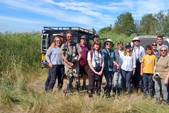 A group of adults and a boy stand in sunshine in front of 4x4 vehicles, with reeds and trees behind