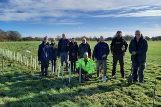 A goroup of people standing in a field alongside a row of planted trees, smiling at the camera