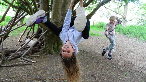 Child smiling to camera, hanging upside down from tree branch with long hair dangling. Another child sits on a rope swing looking on.  