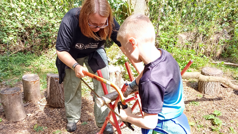 Rebekah leans over a saw horse with a young boy on the other side. They each have a hand on the bow saw as they cut a branch.