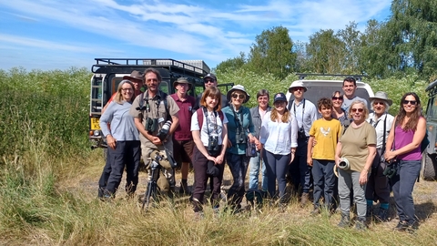 A group of adults and a boy stand in sunshine in front of 4x4 vehicles, with reeds and trees behind