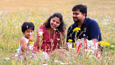 Indian family of mother, father and daughter sit enjoying a picnic in a wildflower meadow. The child is smelling an oxeye daisy. Everyone is smiling