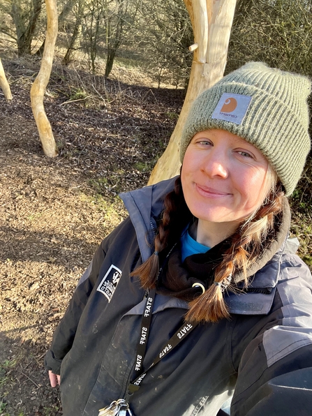 A woman in a sunny woodland with plaited hair wears a pale green beanie and smiles at the camera