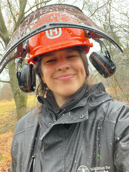 A woman wearing an orange hardhat and ear protectors, with the visor tilted up, smiles at the camera