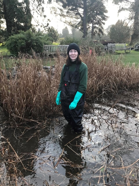 A woman in waders and bright blue rubber gloves stand knee-deep in a pond with a big grin on her face