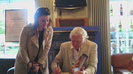 A woman with long dark hair wearing a beige trench coat stands next to a seated Sir David Attenborough as he signs a book