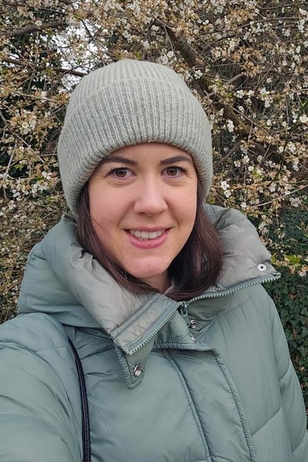 A woman with dark brown hair wearing a grey beanie and pale blue puffer jacket smiles at the camera beside a blossom-covered hedge