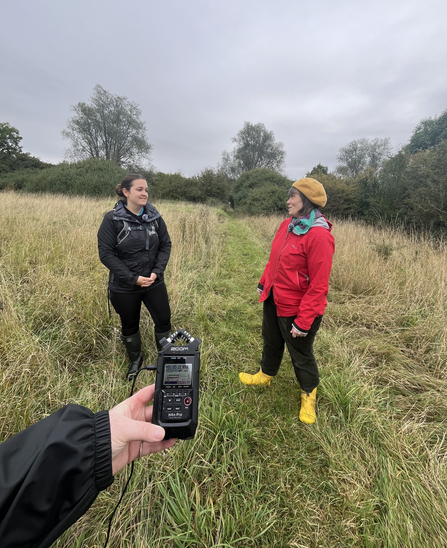 Two women, one dressed in black and the other in red and yellow, stand while talking, an audio recording device held by the photographer in the foreground