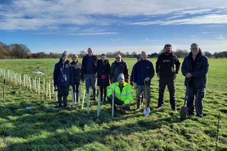 A goroup of people standing in a field alongside a row of planted trees, smiling at the camera